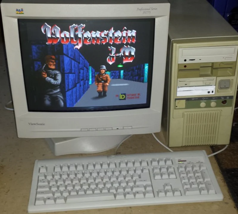 Béžové PC s CRT monitorem, na obrazovce úvodní obrazovka hry Wolfenstein 3D